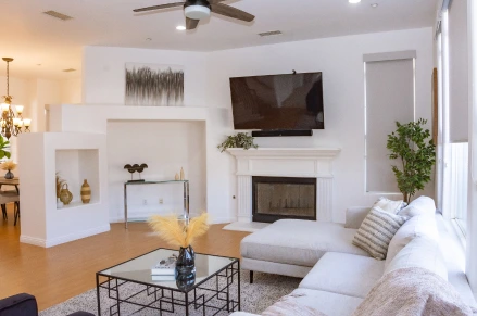 Indoor shot of a bright and airy living room. The room features a light sectional sofa with pillows. A glass coffee table with a black metal frame sits centrally in the room. A fireplace with a white mantelpiece is a focal point on one wall; a large flat-screen television mounted above it.