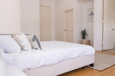 Full indoor shot of a bedroom. A white queen-sized bed is the central focus. The bed's frame is white, and the bedding is a crisp white, with decorative pillows. The overall impression is one of a simple, clean, and bright bedroom design with a calming color palette.
