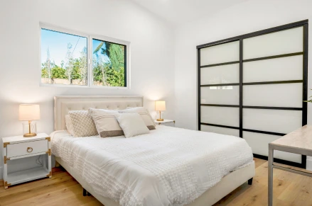 Indoor shot of a bedroom. A light cream-colored bed with a tufted headboard is centered in the image. A pair of sliding doors with a frosted, translucent glass panel. The floor is light oak-colored hardwood planks.
