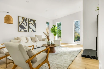 Indoor shot of a modern living room. The room features a light sectional sofa, two matching cream-colored armchairs with wooden frames. A light rug covers the flooring.