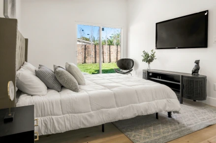 Indoor shot of a bedroom. A queen-sized bed, dressed in white bedding, is centrally located. A flat-screen TV is mounted above the media cabinet, featuring a dark screen.